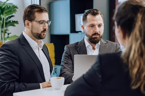 Three people in business attire having a discussion around a laptop at a white table, with coffee and water bottles present.