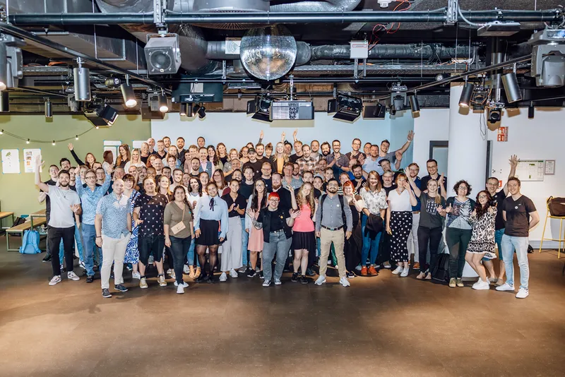 A large group of people posing together indoors, smiling and waving at the camera. The setting appears to be a modern office or event space with lighting and a disco ball overhead.