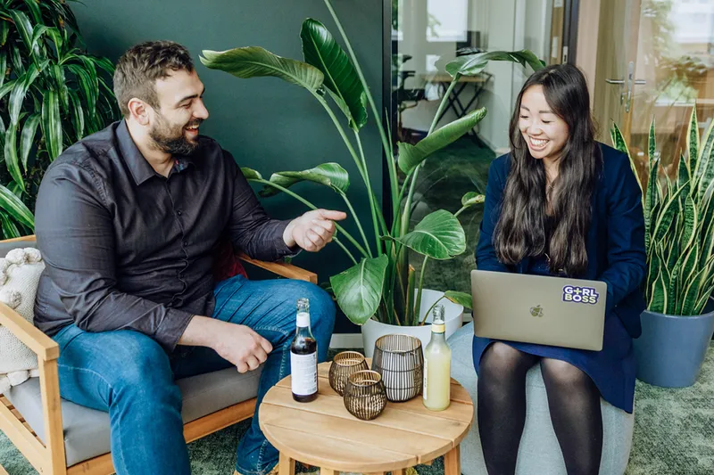 Two people sitting in a modern office space with plants, smiling and talking. One person is holding a laptop with a sticker, and there are drinks on a small table between them.